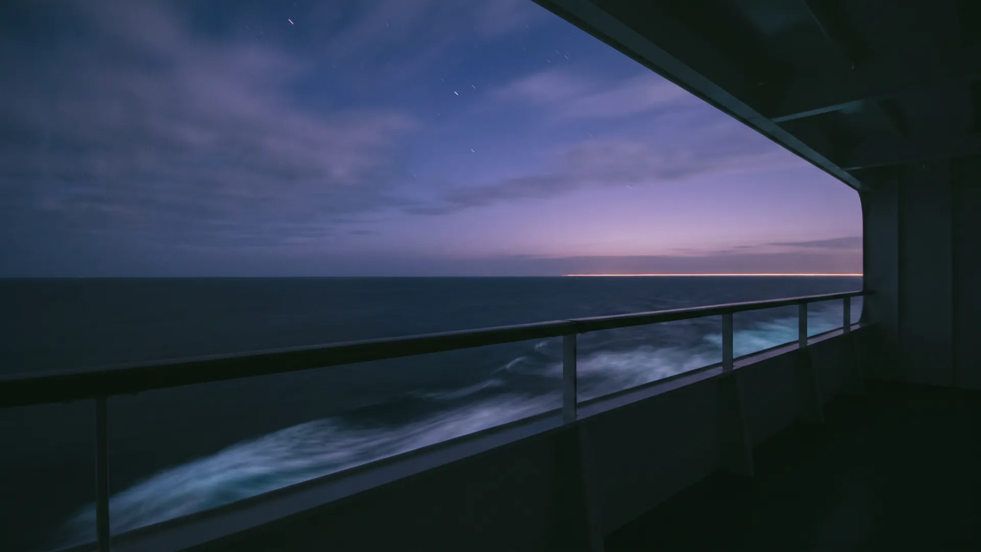 The deck rail of a ship at night, the sea below and a violet afterglow at the horizon.